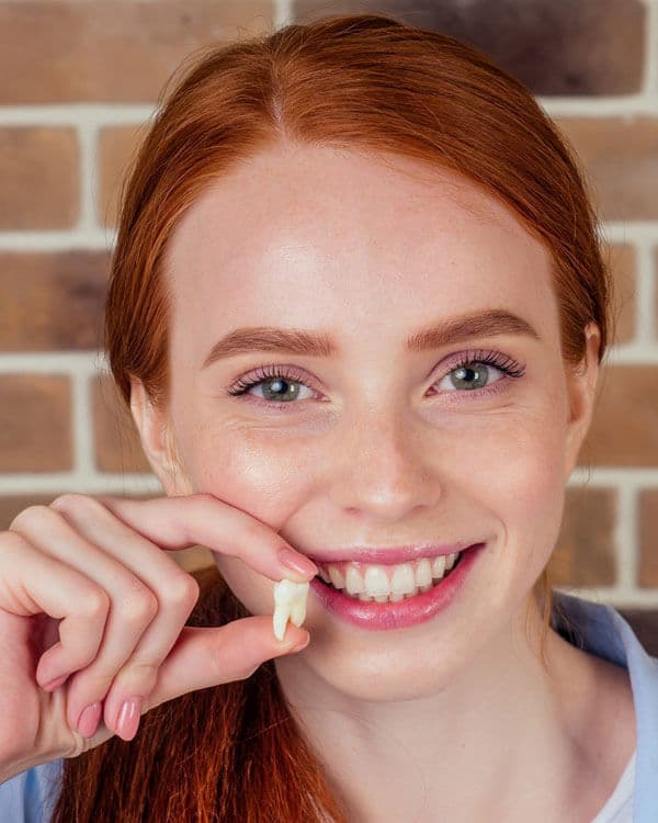Young woman with red hair smiling at camera, finger near mouth, brick wall background.
