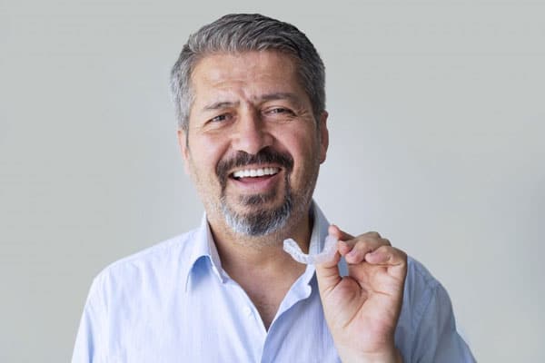 Smiling male dentist in light blue shirt pointing at camera against gray background.