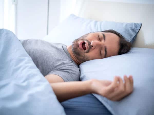 Man sleeping in bed, mouth open, appearing to snore or experience sleep apnea issues.